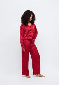 Czerwony satynowy komplet piżamowy składający się z koszuli na guziki z wzorzystym wzorem oraz pasujących szerokich spodni. Tkanina ma gładką, błyszczącą fakturę.