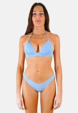 Femme debout, face à l’avant, portant un bikini bleu clair sur un fond uni.
