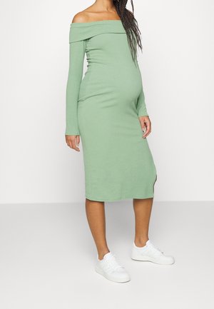 Schwangere Frau, die ein hellgrünes, schulterfreies Midikleid mit langen Ärmeln und weißen Sneakern trägt, stehend vor einem schlichten Hintergrund.