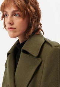 Ung person med kort rött hår bär en olivgrön ullkappa med breda slag och en svart topp, tittar något åt vänster mot en vit bakgrund.