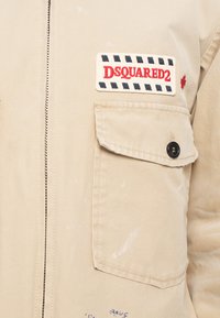 Μπεζ jacket με μπροστινό φερμουάρ, τσέπη με καπάκι και κουμπί, και ραμμένο patch "DSQUARED2" πάνω από την τσέπη.