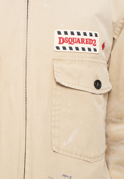 Jachetă bej cu fermoar frontal, buzunar cu clapetă și buton, și o etichetă brodată "DSQUARED2" deasupra buzunarului.