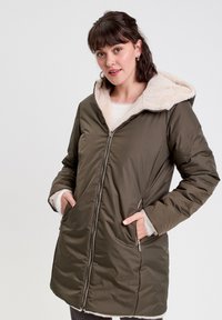 Manteau matelassé vert olive avec une capuche et des poignets en fausse fourrure, deux poches avant et une fermeture éclair centrale. Texture douce et design chaud.