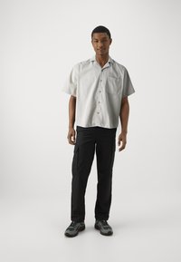 Kurzärmliges, hellgraues knopfbares Hemd mit einer Tasche, kombiniert mit schwarzen Cargo-Hosen. Modell trägt graue Sportschuhe.