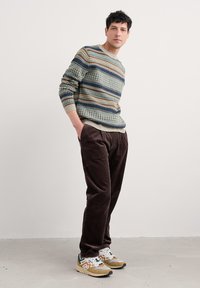Strickpullover mit grünen, blauen und orangefarbenen Streifen; braune Cordhosen; mehrfarbige Turnschuhe mit schwarzen, braunen und weißen Akzenten.