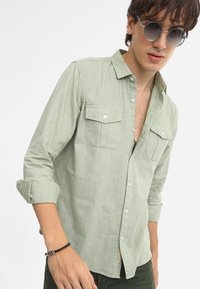 Camisa verde clara de botones hecha de tela texturizada, con dos bolsillos en el pecho y mangas remangadas, combinada con gafas de sol redondas.