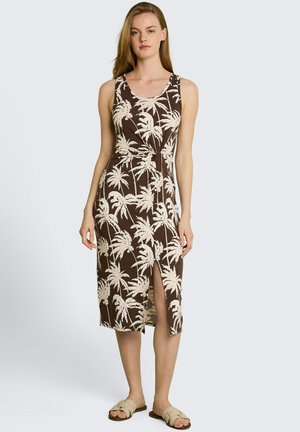 Frau, die ein ärmelloses braunes Midikleid mit beigem Palmenmuster und Frontschlitz trägt, kombiniert mit beigen flachen Sandalen.