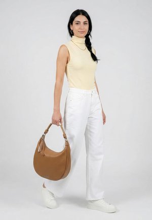 Frau mit langem schwarzem Zopf, die einen ärmellosen beigen Rollkragenpullover und weiße Hosen trägt, hält eine große braune Handtasche und weiße Sneaker.