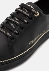 Černé kožené tenisky s logem Emporio Armani na šňůrkách, po straně a na kovových očkách, s texturovanou černou podrážkou a bílým lemováním.