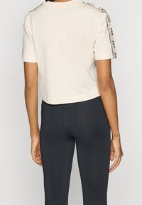 Osoba ubrana w białą, krótką koszulkę z wzorzystymi paskami na rękawach i czarne dopasowane legginsy, pokazująca widok z tyłu oraz tatuaż na ramieniu.