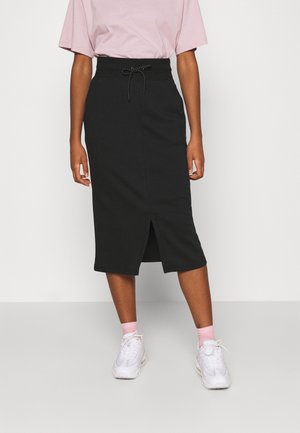 Vrouw die een zwarte knielange rok met een split aan de voorkant draagt, een roze T-shirt, lichtroze sokken en witte sneakers, staand op een lichtgrijze vloer.