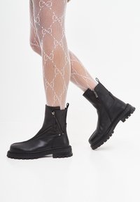 Botines de cuero negro con suela gruesa, cremalleras laterales y textura suave, usados con pantimedias de red blanca con patrón.