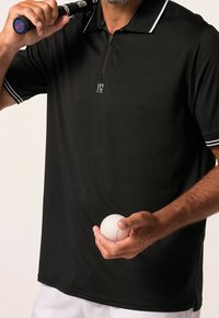 Polo noir à manches courtes, col zippé et accents blancs. Tenant une balle blanche dans une main, avec une raquette posée sur l'épaule opposée.