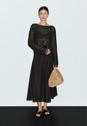 Black crocheted long-sleeve top with open weave, paired with a flowing black skirt. Knotted necklace, tan suede bag, and dark sandals.