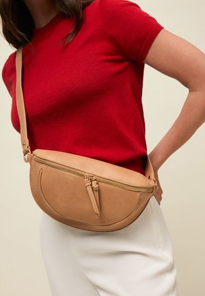 Mujer que lleva una camiseta roja de manga corta y pantalones blancos con un bolso bandolera de cuero marrón claro con un bolsillo frontal curvado con cremallera.