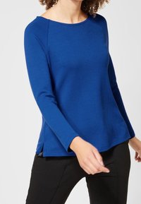 Blå långärmad tröja med rund halsringning, sidofickor och mjukt material, i kombination med svarta byxor.