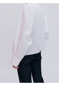 Chemise blanche en coton à manches bouffantes, poignets boutonnés et coupe décontractée, associée à un jean en denim foncé. Design minimaliste et texture lisse.