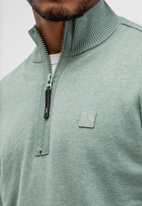 Man in een lichtgroen sweater met kwartrits en een geribde kraag, met een klein geborduurd logo op de borst, over een wit onderhemd.