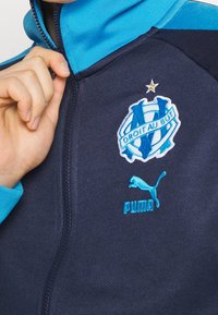 Marinblå zip-up jacka med turkosa accenter, broderad Olympique de Marseille-logotyp och Puma-logotyp på nedre högra sidan. Slät textur.