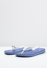 Modré žabky s bílými popruhy, strukturovanou stélkou a zeleným logem. Podešev má kontrastní žlutý lem.