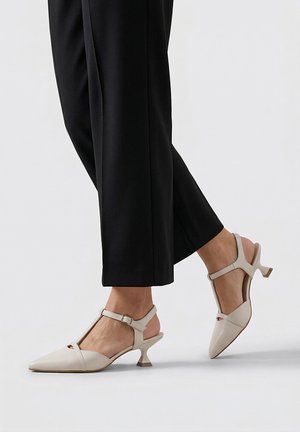 Pies con zapatos de tacón kitten de cuero beige con tira en T, combinados con pantalones negros de corte recto sobre un fondo liso.