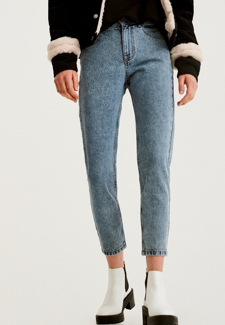 Personne portant un jean bleu clair taille haute à jambes droites, un haut noir, une veste noire avec des poignets en fourrure blanche, et des bottines blanches à semelles épaisses.