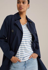 Navy blue trench coat with double-breasted buttons, worn over a white and blue striped v-neck shirt, paired with light jeans.