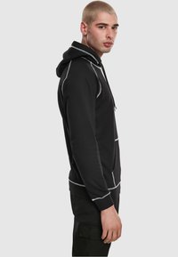 Crna dukserica s kapuljačom i bijelim ukrasnim šavovima, s vrpcom za vezanje kapuljače i bočnim džepovima. Mekani materijal s opuštenim krojem i rebrastim manšetama.