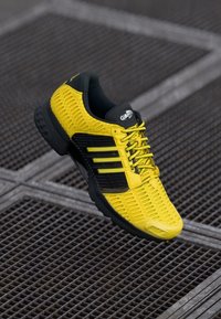 Žlutá sportovní bota s texturovaným svrškem, černými detaily a třemi pruhy. Obsahuje polstrovanou podešev a reflexní prvky.