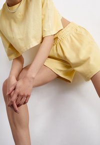 Person som bär en ljusgul kortärmad t-shirt och elastiska shorts, poserad med böjda knän och händerna knäppta nära ett ben, mot en vit bakgrund.