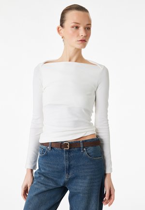 Vrouw met een wit, langmouwen, boothals shirt dat in een blauwe spijkerbroek met een bruine riem is gestopt, kijkend naar de zijkant tegen een effen achtergrond.