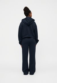 Κοντό φούτερ σε ναυτικό μπλε με κουκούλα, σε συνδυασμό με παντελόνι φαρδιάς γραμμής, κατασκευασμένο από απαλό ύφασμα. Απλός σχεδιασμός χωρίς ορατά μοτίβα ή διακοσμητικά στοιχεία.