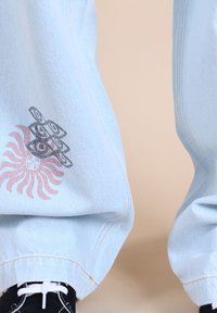 Pantalons en denim oversize bleu clair avec imprimé graphique présentant un soleil rose et des formes géométriques noires. Coutures blanches et coupe décontractée.