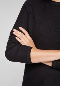 Pull en maille noire avec un motif texturé, doté d'un col rond et de manches longues à poignets. Les bras sont croisés devant le corps.
