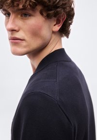 Νέος άνδρας με καστανά σγουρά μαλλιά, που φοράει σκούρο μπλε πουλόβερ, κοιτάζει πλάγι α σε απλό φωτεινό φόντο.