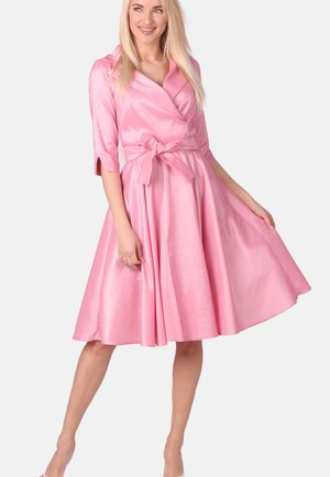 Frau lächelt, trägt ein glänzendes pinkfarbenes knielanges Kleid mit dreiviertel Ärmeln und einer gebundenen Taillenschleife, hält den Rock leicht ausgestreckt.