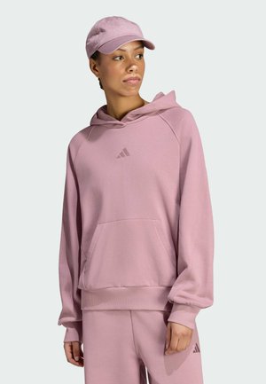 Vrouw draagt een roze Adidas hoodie, bijpassende broek en een roze pet, kijkt iets naar de zijkant tegen een effen achtergrond.