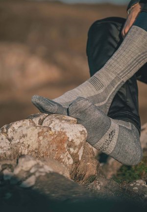 Pieds portant des chaussettes d'extérieur grises reposant sur un terrain rocheux, avec une personne vêtue d'un pantalon noir, en plein air sous la lumière naturelle.