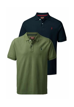 Uomo con polo blu navy, jeans blu e stivali marroni in piedi contro uno sfondo bianco uniforme, che guarda verso il basso sorridendo.