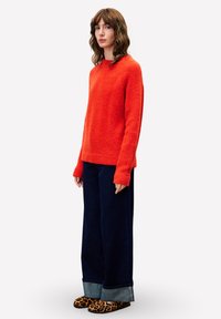 Frau mit schulterlangem braunen Haar, die einen leuchtend orangefarbenen Pullover, weite, dunkle Jeans mit Umschlägen und Schuhe mit Leopardenmuster trägt und seitlich steht.