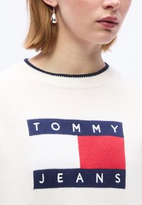 Κοντινό πλάνο ατόμου που φοράει λευκό πουλόβερ με λογότυπο Tommy Jeans σε μπλε και κόκκινο και ασημένιο σκουλαρίκι σε σχήμα δάκρυ.