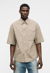 Hombre con camisa de manga corta de cuadros beige y marrón con bolsillo frontal, combinado con jeans azul claro, posando frente a un fondo blanco.
