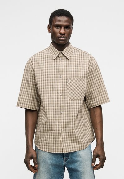 Homme portant une chemise à manches courtes à carreaux beige et marron avec une poche avant, associée à un jean bleu clair, debout devant un fond blanc.