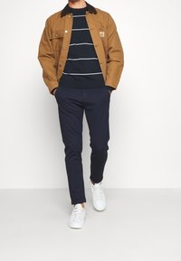 Veste en toile marron avec un col en velours côtelé noir, pull rayé bleu marine, pantalon bleu marine et baskets blanches. Considéré comme une tenue décontractée.