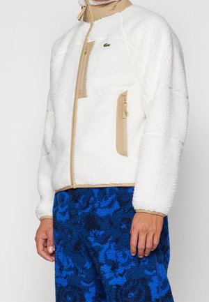 Uomo che indossa una giacca in pile bianca con rifiniture beige e pantaloni in pile blu con motivo, in piedi davanti a uno sfondo semplice.