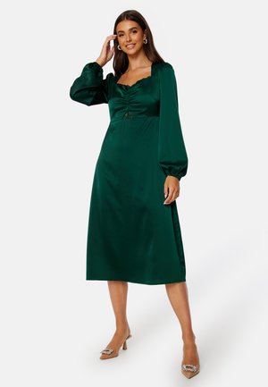 Emerald groene satijnen midi-jurk met lange pofmouwen, vierkante halslijn, geplooid lijfje en kantdetails op de borst. Hakken met embellissementen.