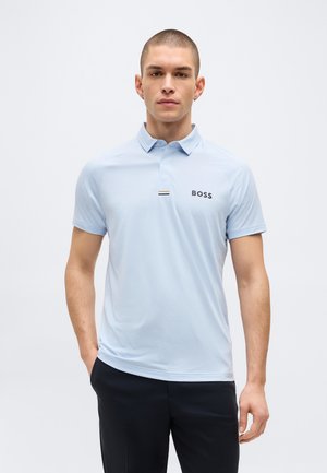 Muž v svetlomodrom polotričku BOSS a čiernych nohaviciach stojí pred čistým bielym pozadím s jednou rukou v vrecku.