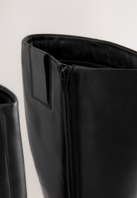 Bottes en cuir noir à texture lisse ; disposent d'une fermeture éclair latérale et d'un haut arrondi, conçues pour une silhouette ajustée.