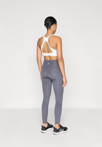 Brassière de sport blanche avec un dos découpé, associée à un legging gris ajusté. Le modèle porte des chaussures de sport noires avec des semelles blanches et trois bandes.