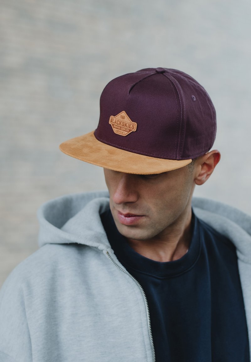 Bordeaux muts met een tan suède klep, met een leren patch waarop de tekst "BLACKSKIES" staat. Gdragend met een grijze hoodie, tegen een vervaagde achtergrond.